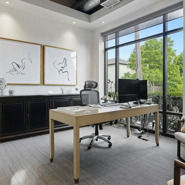 Oficina moderna con escritorio, silla, TV y grandes ventanales con vistas al exterior.